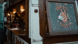 A food truck with wooden accents and a window displaying a sign that reads 'Mean Dogs' accompanied by a cartoon image of a fox. The interior of the truck is warmly lit by small hanging lamps, and there are various items such as containers and possibly food preparation tools visible inside.