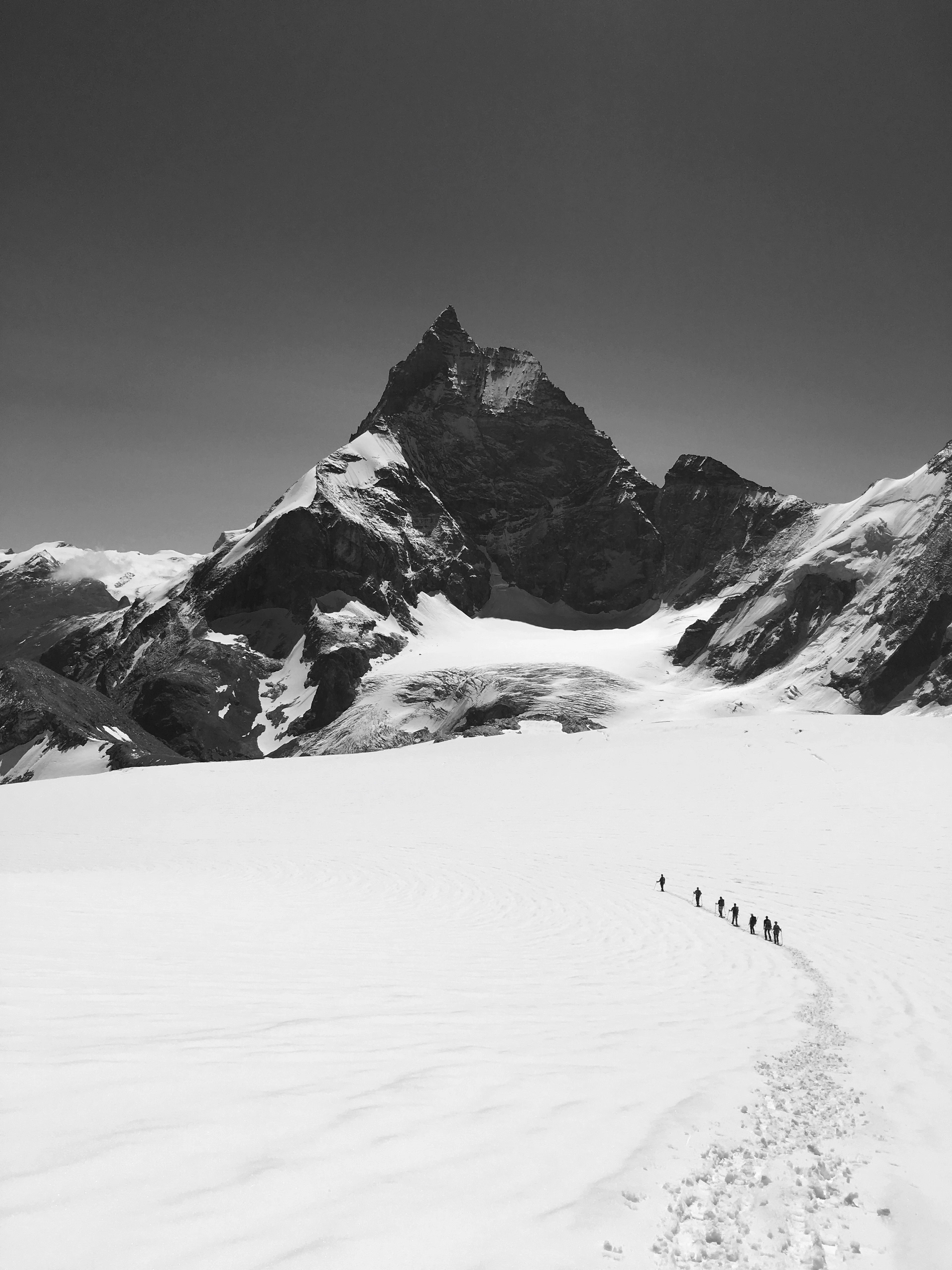 A black-and-white shot of roped climbers crossing a snowfield toward a sharp rocky summit.