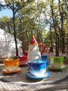A large teapot and teacups are part of an amusement ride nestled among trees. Each cup is vibrantly colored, and the setup is on a wooden platform with a white fence in the background.