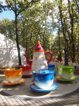 A large teapot and teacups are part of an amusement ride nestled among trees. Each cup is vibrantly colored, and the setup is on a wooden platform with a white fence in the background.