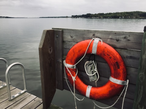 An orange lifebuoy is mounted on a wooden panel by the edge of a dock, overlooking a calm and expansive body of water. The sky is overcast, creating a serene and tranquil atmosphere. A rescue rope is attached to the lifebuoy, ready for use in case of an emergency.