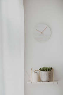 A minimalistic room decor featuring a small round marble wall clock with wooden hands, a white curtain partially covering one side, and a small shelf. The shelf displays a white watering can with a brass handle and a potted succulent plant in a textured concrete pot.