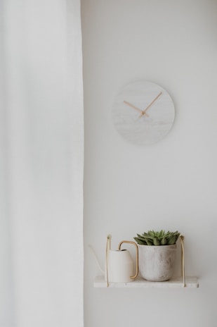 A minimalistic room decor featuring a small round marble wall clock with wooden hands, a white curtain partially covering one side, and a small shelf. The shelf displays a white watering can with a brass handle and a potted succulent plant in a textured concrete pot.