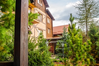 A rustic wooden house with a red-tiled roof surrounded by lush green pine trees and foliage. The exterior features large windows and wooden paneling. The atmosphere is serene and natural.