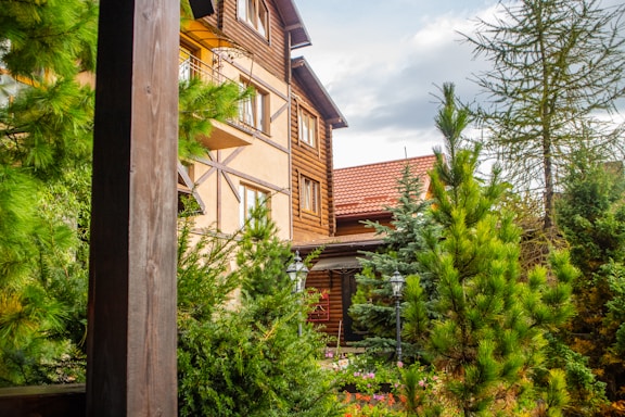 A rustic wooden house with a red-tiled roof surrounded by lush green pine trees and foliage. The exterior features large windows and wooden paneling. The atmosphere is serene and natural.
