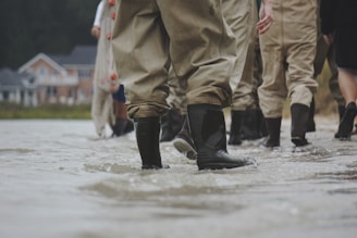 Several people wearing waterproof trousers and boots walk through shallow water. In the background, there is a blurry view of houses, suggesting a residential area.