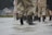 Several people wearing waterproof trousers and boots walk through shallow water. In the background, there is a blurry view of houses, suggesting a residential area.