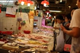 A bustling seafood market with people examining a variety of fresh fish and seafood displayed on a long counter. The setting features overhead red lamps casting a warm glow, creating a lively and busy atmosphere. There are baskets and signs on the walls, with tiles showing signs of wear.