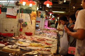 A bustling seafood market with people examining a variety of fresh fish and seafood displayed on a long counter. The setting features overhead red lamps casting a warm glow, creating a lively and busy atmosphere. There are baskets and signs on the walls, with tiles showing signs of wear.