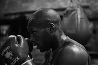 A person wearing boxing gloves in a gym is captured in a focused stance with a punching bag in the background. The scene is in black and white, highlighting the intense concentration of the individual as they prepare to box.