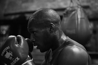 A person wearing boxing gloves in a gym is captured in a focused stance with a punching bag in the background. The scene is in black and white, highlighting the intense concentration of the individual as they prepare to box.