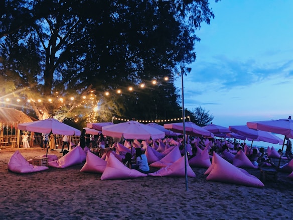 A beach scene featuring numerous pink bean bags and umbrellas arranged on the sand. There are fairy lights strung across the area, creating a warm glow against the evening sky. A few people are gathered in small groups, sitting or standing by tables made of wooden pallets. Tall trees and the ocean are visible in the background, with a serene blue sky above.
