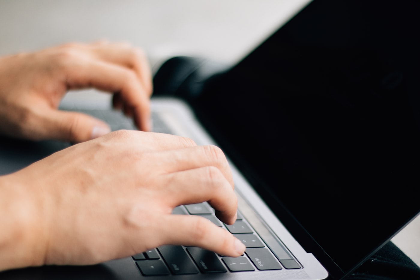 Close-up photo of hands typing on a keyboard with code visible on a blurred background screen.