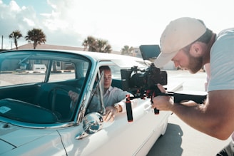 A man is inside a vintage car while another man outside is filming him using a professional camera mounted on a stabilizer. The scene takes place in a sunny outdoor setting with palm trees and buildings in the background.