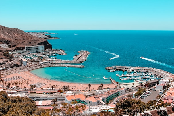 A stunning coastal view reveals a vibrant seaside landscape. A wide expanse of blue ocean meets a sandy beach, populated with organized rows of sunbeds and umbrellas. Adjacent to the beach, a marina is filled with numerous boats docked in neat rows. Several modern buildings and hotels are nestled against the hillside, offering scenic views of the water. Lush palm trees are scattered throughout the area, adding to the tropical atmosphere.