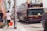 A double-decker tour bus with a maroon and beige color scheme is parked on a busy street. Several people are visible on the upper deck of the bus. In the foreground, a person holding an orange bucket is walking alongside the bus. The street is lined with murals and posters, and there is a palm tree in the background.