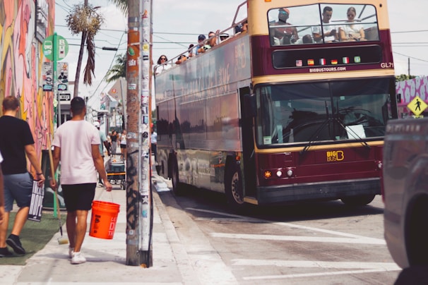 A double-decker tour bus with a maroon and beige color scheme is parked on a busy street. Several people are visible on the upper deck of the bus. In the foreground, a person holding an orange bucket is walking alongside the bus. The street is lined with murals and posters, and there is a palm tree in the background.