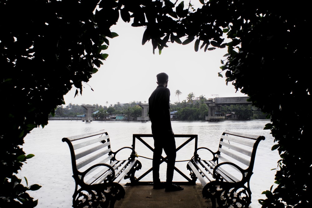 man standing near outdoor bench facing on body of water viewing houses and buildings during daytime