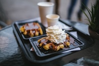 A black tray holds two servings of waffles covered in chocolate sauce, with a generous dollop of whipped cream on one. Two white paper cups are placed next to them on a clear glass-top table. In the background, there is a blurred view of greenery.