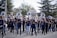 A marching band is performing outdoors, with a group of musicians playing instruments like trombones and saxophones. They are wearing blue uniforms with matching logos, and are marching in formation on a street. The background shows spectators and trees.