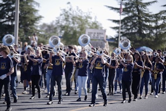 A marching band is performing outdoors, with a group of musicians playing instruments like trombones and saxophones. They are wearing blue uniforms with matching logos, and are marching in formation on a street. The background shows spectators and trees.