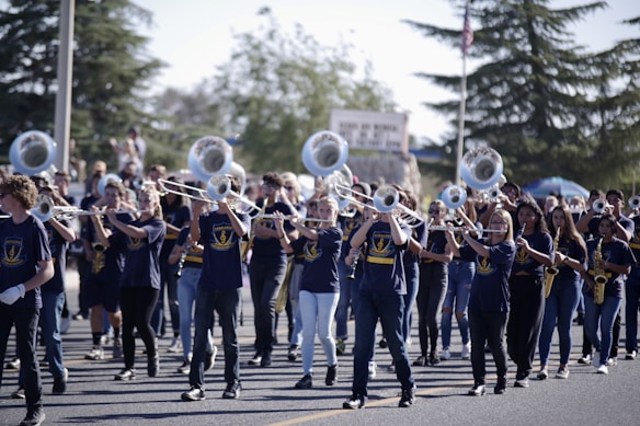 A marching band is performing outdoors, with a group of musicians playing instruments like trombones and saxophones. They are wearing blue uniforms with matching logos, and are marching in formation on a street. The background shows spectators and trees.