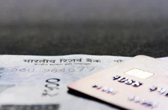A close-up view of a credit card positioned on top of an Indian currency note, with a visible microchip and embossed numbers on the card. The focus is on both financial items, showcasing the intricacy of details like the microchip and textual elements.