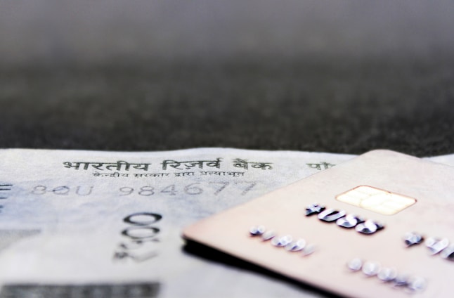 A close-up view of a credit card positioned on top of an Indian currency note, with a visible microchip and embossed numbers on the card. The focus is on both financial items, showcasing the intricacy of details like the microchip and textual elements.