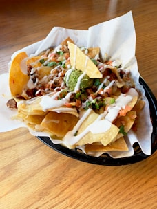 A plate of nachos topped with melted cheese, diced tomatoes, cilantro, guacamole, and drizzled with white sauce. The dish is served on a black plate lined with parchment paper atop a wooden table.