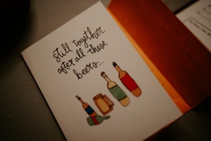 A greeting card with an illustration of beer bottles and mugs, accompanied by the handwritten text 'Still together after all those beers...'. The card is partially open, revealing an orange interior and additional handwritten notes inside.