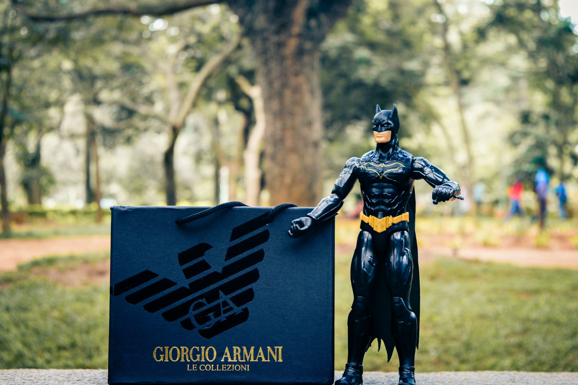 A toy figure resembling a superhero is standing on a stone surface in an outdoor setting. Next to the figure is a shopping bag with a designer logo and brand name written on it. The background features blurred trees and a path, suggesting a park-like environment.
