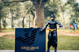 A toy figure resembling a superhero is standing on a stone surface in an outdoor setting. Next to the figure is a shopping bag with a designer logo and brand name written on it. The background features blurred trees and a path, suggesting a park-like environment.