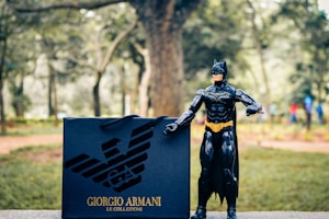 A toy figure resembling a superhero is standing on a stone surface in an outdoor setting. Next to the figure is a shopping bag with a designer logo and brand name written on it. The background features blurred trees and a path, suggesting a park-like environment.