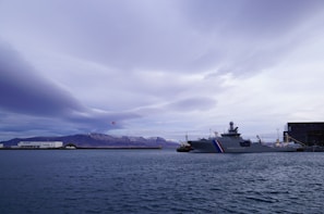 A large coastal patrol ship is docked in a harbor with a mountainous landscape in the background. Above the mountains, clouds fill the sky, and a helicopter is visible in mid-flight. The water in the foreground is calm, creating a serene atmosphere.