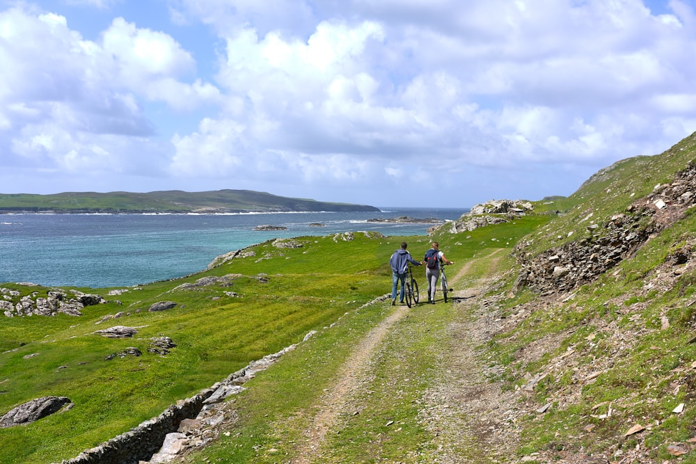 Deux personnes marchant ensemble avec des vélos dans un champ vert regardant la montagne et la mer bleue sous le ciel blanc et bleu pendant la journée