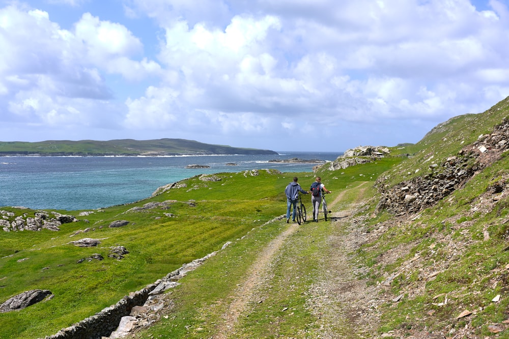 zwei Menschen, die zusammen mit Fahrrädern auf der grünen Wiese gehen und tagsüber Berge und blaues Meer unter weißem und blauem Himmel betrachten