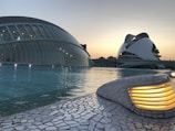 A futuristic architectural scene with a sweeping glass dome structure adjacent to a uniquely designed white building. The scenery is set against a serene evening sky, with reflections on the tranquil water surrounding the buildings. A lit stone path curves along the edge, adding subtle illumination.