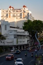 A city street scene featuring a multi-story white building with red roof accents and a sign reading 'BEKIZAAR HOTEL'. The street below is bustling with traffic, including cars and motorbikes. The environment includes green trees and decorative sculptures along the roadside. The sunlight creates shadows and highlights the architecture.