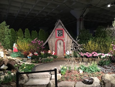 A whimsical garden scene features a small, rustic wooden house with decorative red trim, surrounded by vibrant flowers and lush greenery. Cutout figures of cartoon characters are posed amidst the garden beds, adding a playful, storybook element to the setting. Stone pathways and decorative elements like mushrooms enhance the fairy tale atmosphere.