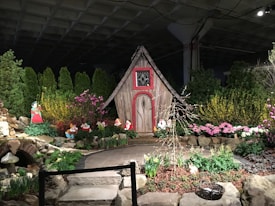 A whimsical garden scene features a small, rustic wooden house with decorative red trim, surrounded by vibrant flowers and lush greenery. Cutout figures of cartoon characters are posed amidst the garden beds, adding a playful, storybook element to the setting. Stone pathways and decorative elements like mushrooms enhance the fairy tale atmosphere.