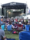 A large outdoor music festival features a stage with a band performing and a crowd of people gathered in front on a grassy area. Attendees are seated on portable chairs or standing as they watch the performance. The atmosphere is lively with stage lighting and a large screen in the background.