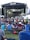A large outdoor music festival features a stage with a band performing and a crowd of people gathered in front on a grassy area. Attendees are seated on portable chairs or standing as they watch the performance. The atmosphere is lively with stage lighting and a large screen in the background.