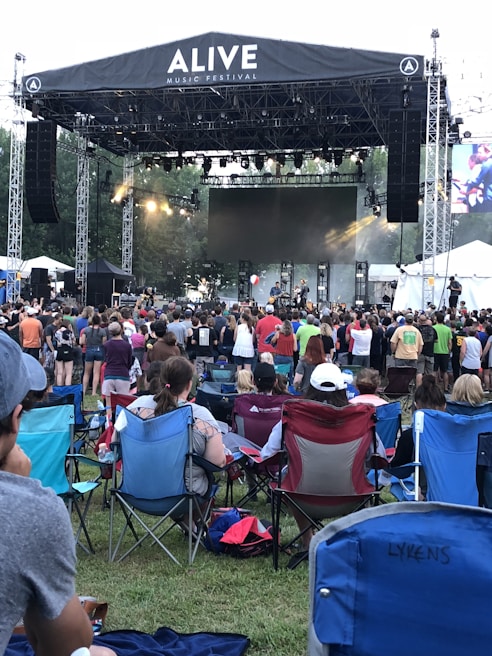 A large outdoor music festival features a stage with a band performing and a crowd of people gathered in front on a grassy area. Attendees are seated on portable chairs or standing as they watch the performance. The atmosphere is lively with stage lighting and a large screen in the background.