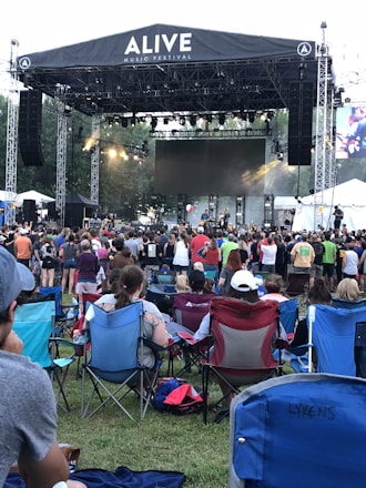 A large outdoor music festival features a stage with a band performing and a crowd of people gathered in front on a grassy area. Attendees are seated on portable chairs or standing as they watch the performance. The atmosphere is lively with stage lighting and a large screen in the background.