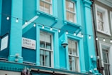 A bright blue building with large windows is decorated with a string of hanging lights. A street sign reads 'Kingly Street W1, City of Westminster' against the vibrant background. The architecture features classic elements, and there are floral decorations visible.