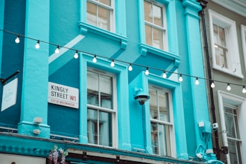 A bright blue building with large windows is decorated with a string of hanging lights. A street sign reads 'Kingly Street W1, City of Westminster' against the vibrant background. The architecture features classic elements, and there are floral decorations visible.