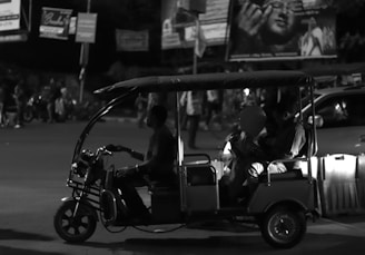 A black-and-white image depicting a three-wheeled vehicle, similar to a tuk-tuk, with a driver and passengers. The passengers seem to be holding balloons. The scene is set on a street at night with blurred pedestrians and vehicles in the background.