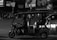 A black-and-white image depicting a three-wheeled vehicle, similar to a tuk-tuk, with a driver and passengers. The passengers seem to be holding balloons. The scene is set on a street at night with blurred pedestrians and vehicles in the background.