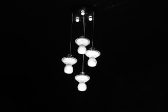 A group of ornate, glass chandeliers hang from the ceiling, illuminated against a dark background. The light fixtures have a vintage design, each consisting of multiple glass sections that reflect light.