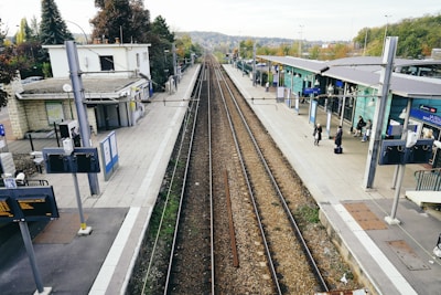A railway station platform featuring train tracks running down the center, bordered by platforms on either side. The area appears relatively quiet with a few people waiting. Buildings and signage typical of a train station are visible, along with a small number of trees and greenery in the background.